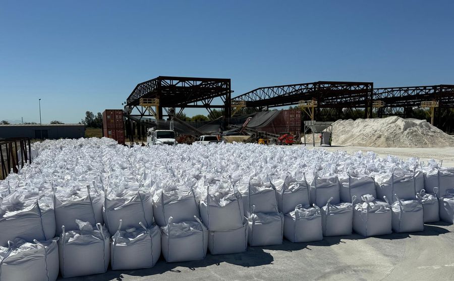 Rows of filled white bulk bags stacked two high in the yard, with steel structures and a stockpile behind