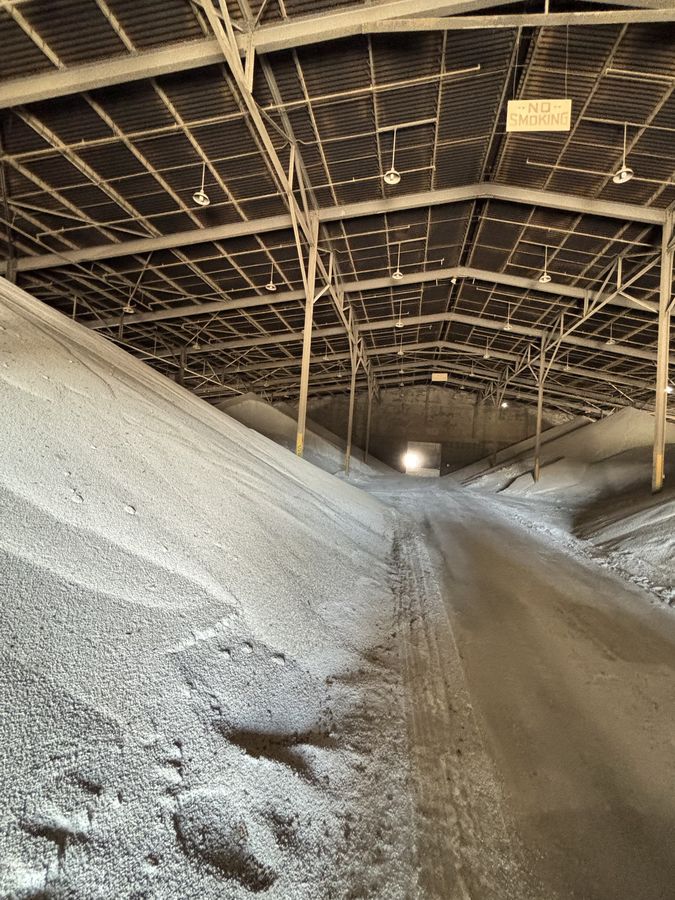Inside a long steel-framed storage building, granular material banked high on both sides of a central lane