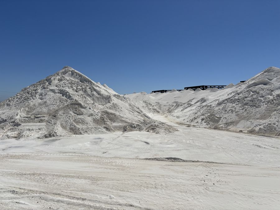 Two large white aragonite stockpiles under a clear blue sky, with steel structures on the skyline between them